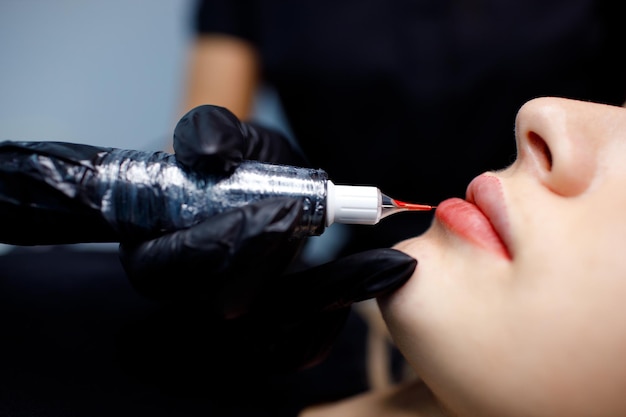 Cosmetologista aplicando tatuagem de maquiagem permanente vermelha nos lábios de mulher jovem Conceito de procedimento de tatuagem de lábios permanentes