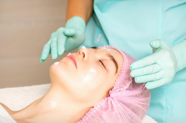 Cosmetologista com mãos enluvadas aplica uma máscara hidratante com creme peeling no rosto feminino Tratamento de cosmetologia facial Procedimentos para cuidados faciais
