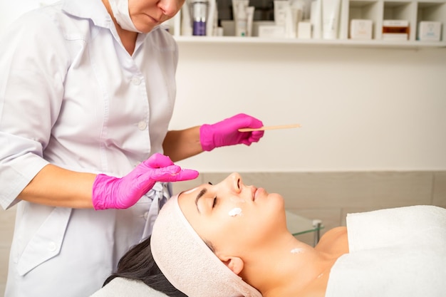 Cosmetologista com mãos enluvadas aplica uma máscara hidratante com creme peeling no rosto feminino Tratamento de cosmetologia facial Procedimentos para cuidados faciais