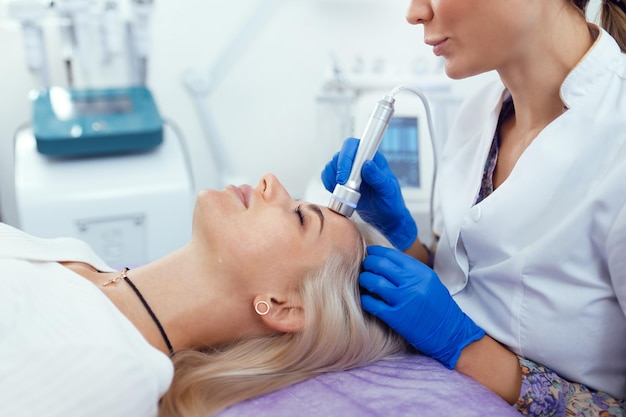 Cosmetologista fazendo procedimento de facail de fonoforese de aparelho para mulher em salão de beleza