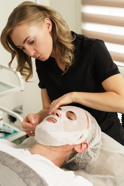 Cosmetologista feminina aplicando máscara de folha no rosto do cliente na clínica de beleza