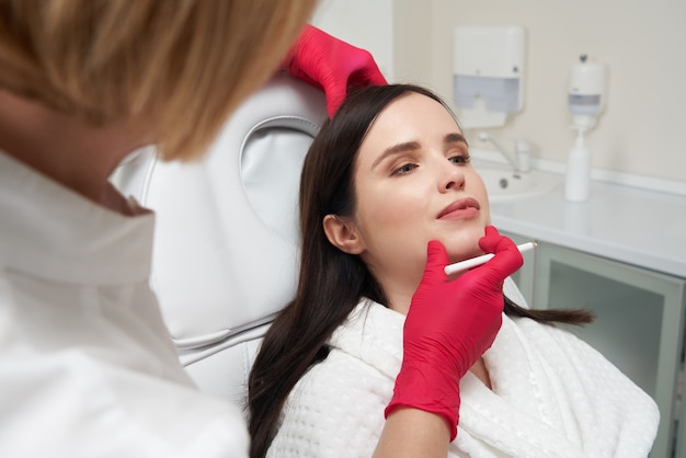 Cosmetologista feminina desenhando uma linha no rosto feminino com um marcador branco para se preparar para o procedimento