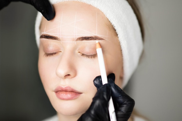 Cosmetologista mapeando sobrancelhas de mulher com lápis branco antes do tratamento de maquiagem permanente