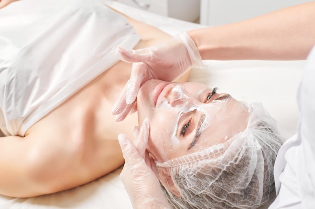 Cosmetologista massageia máscara de creme na pele do rosto de mulher para procedimento de rejuvenescimento em salão de beleza