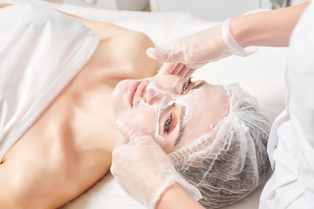 Cosmetologista massageia máscara de creme na pele do rosto de mulher para procedimento de rejuvenescimento em salão de beleza