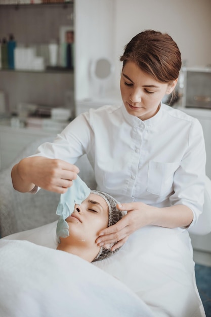 Cosmetologista remove a máscara de creme branco no rosto de uma mulher caucasiana que está deitada no sofá ...