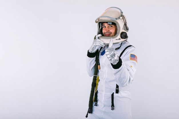 Cosmonauta masculino en traje espacial y casco, sosteniendo el casco con las manos y sonriendo, sobre fondo blanco.