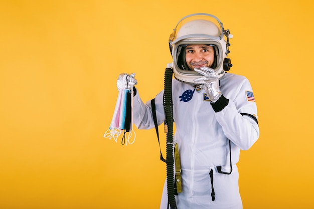 Cosmonauta masculino en traje espacial y casco, sosteniendo muchas máscaras quirúrgicas de colores, en la pared amarilla. Covid19 y concepto de virus