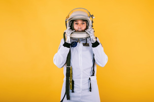 Cosmonauta masculino en traje espacial y casco, sosteniendo su casco con las manos con gesto de sorpresa, en la pared amarilla.