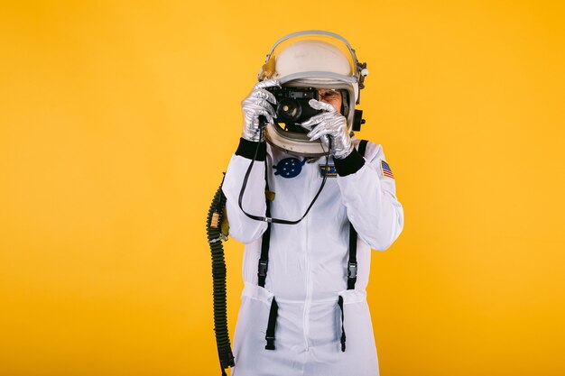 Cosmonauta masculino en traje espacial y casco, tomando una foto con una cámara retro, en la pared amarilla.