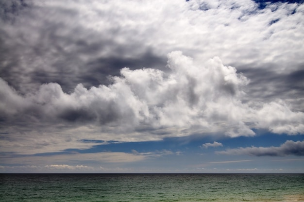 Foto costa do mar e praia rochosa, céu azul com nuvens brancas, montanhas e uma pequena vila ao fundo