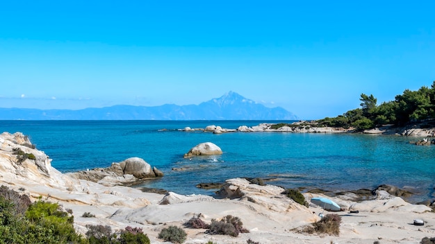 Costa del mar Egeo con gente nadando, rocas sobre el agua y la tierra en la distancia, vegetación, agua azul, Grecia