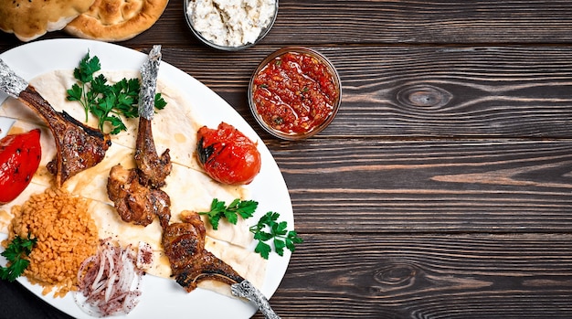 Costeleta de cordeiro ou pirzola kuzu turca com bulgur, legumes grelhados e especiarias em um prato branco. Fundo de madeira escura, vista superior com espaço de cópia