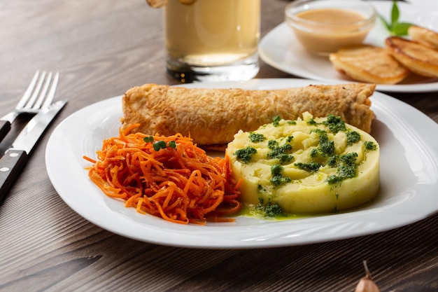 Costeleta de porco crocante servida com purê de batata quente polvilhado com cebola verde e salada de cenoura em um prato branco sobre uma mesa de madeira