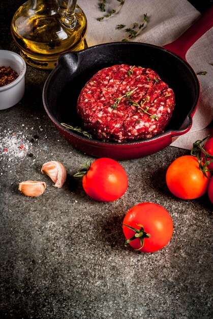 Costeletas de hambúrgueres de carne orgânica crua com especiarias, tomilho, tomate, azeite na frigideira em preto, copyspace