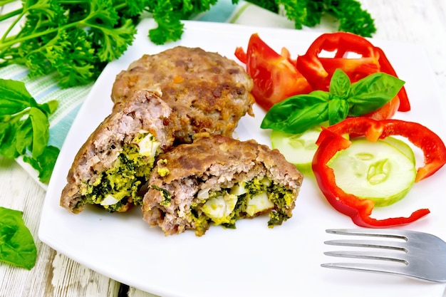 Costeletas recheadas com espinafre e ovo, salada com tomate, pepino e pimenta em um prato sobre uma toalha, manjericão e salsa em um fundo de tábua de madeira