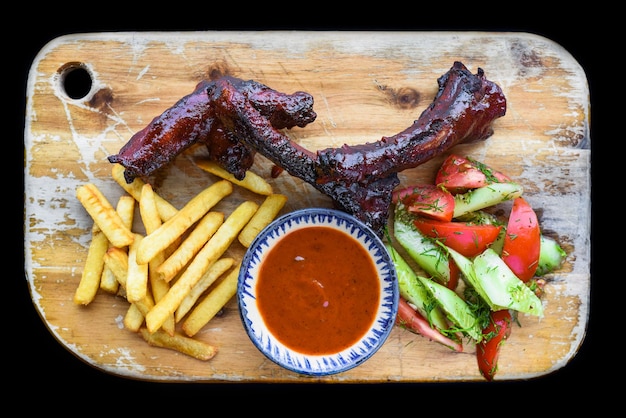 Costelinha de porco defumada grelhada com batatas fritas e molho e salada na tábua de madeira sobre fundo preto t