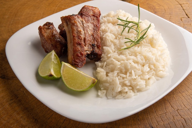 Costelinha de porco frita, arroz e condimentos em um prato branco organizado em madeira rústica clara, foco seletivo.