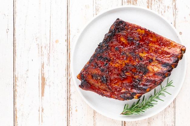 Costillas de cerdo asadas a la barbacoa con miel, orégano y romero en un plato de cerámica simplemente blanco sobre fondo blanco de textura de madera vieja con espacio para copiar texto, vista superior