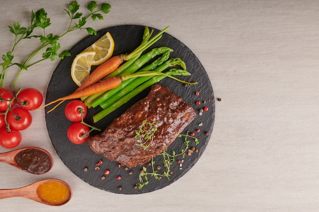 Costillas de cerdo asadas a la parrilla de una barbacoa de verano servidas con verduras, espárragos, zanahorias baby, tomates frescos y especias. costillas ahumadas sobre superficie de piedra negra. vista superior,