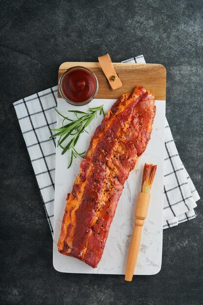 Costillas de cerdo Carne cruda Costillas de cerdo crudas en adobo con especias, salsa de tomate, romero y ajo sobre soporte de mármol blanco sobre fondo de mesa de piedra negra Concepto de barbacoa Vista superior Maqueta