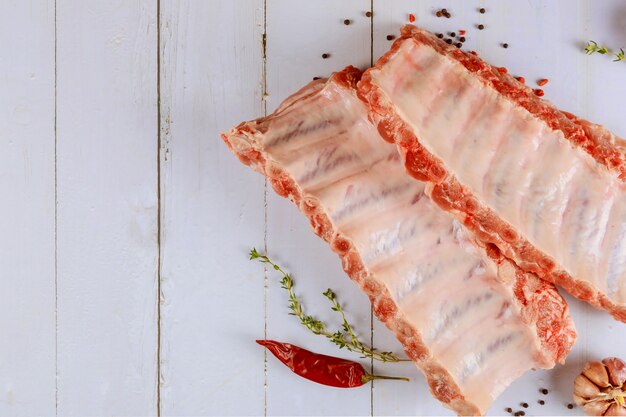 Costillas de cerdo crudas frescas con ingredientes hierbas y especias listas para marinar en una mesa de madera blanca.