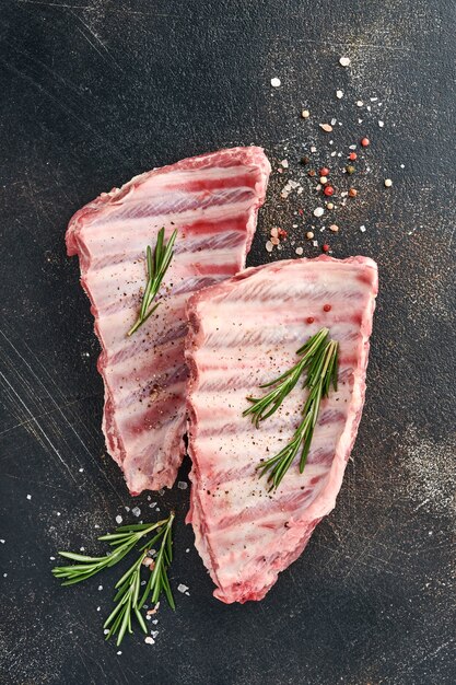 Costillas de cerdo crudas o carne fresca cruda con especias en bandeja de madera negra con pimentón, dientes de ajo y hierbas. Fondo de textura oscura con espacio de copia de texto.