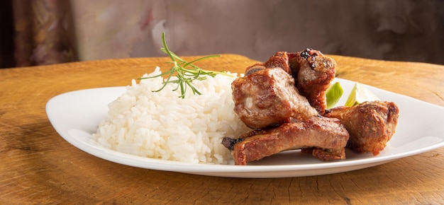 Costillas de cerdo fritas, arroz y condimentos en un plato blanco dispuestas en madera rústica clara, enfoque selectivo.