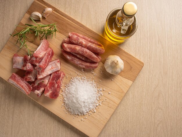 Costillas de cerdo, gajos de salchicha, sal de roca, ajo, cebolla, romero y aceite de oliva sobre una madera rústica clara sobre una mesa, vista superior.