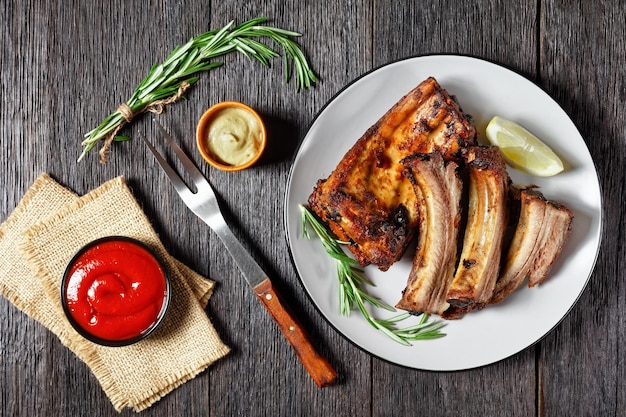 Costillas de cerdo a la parrilla con limón y romero en un plato sobre una mesa de madera oscura con vista horizontal de ketchup y mostaza desde arriba