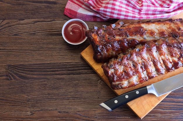 Costillas de cerdo a la parrilla sobre una tabla de cortar de madera con cuchillo de cocina y salsa de tomate sobre fondo de madera marrón. Vista superior, endecha plana.