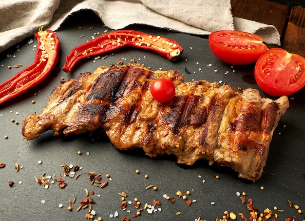 Costillas de cerdo a la parrilla en un tablero negro, tomates rojos frescos y chiles