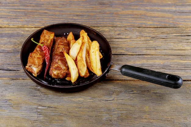 Costillas fritas con las patatas en un sartén en una tabla rústica de madera.