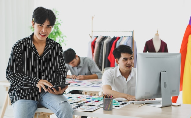 Costureira asiática usando um tablet perto de colegas do sexo masculino, navegando dados no computador e desenhando um esboço