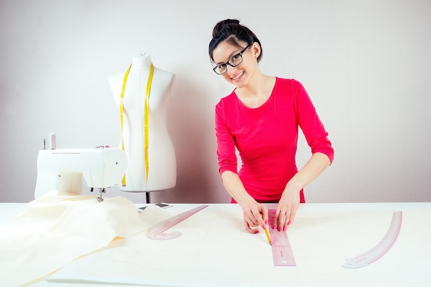 Costureira linda garota com óculos trabalhando com lápis e padrões Fundo de manequim com máquina de costura de fita métrica amarela e fundo branco