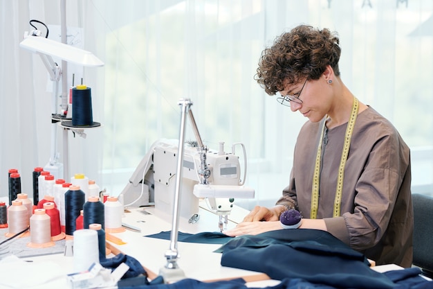 Costureira muito jovem sentada ao lado da máquina de costura e processando tecido azul enquanto trabalhava em uma peça de roupa