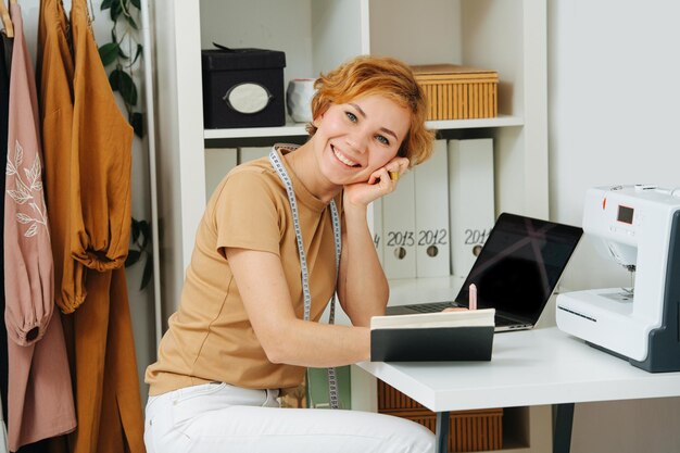 Costureira radiantemente sorridente, sentada atrás de sua mesa de trabalho, escrevendo em um livro-caixa Medidor pendurado no pescoço Prateleiras e prateleiras atrás