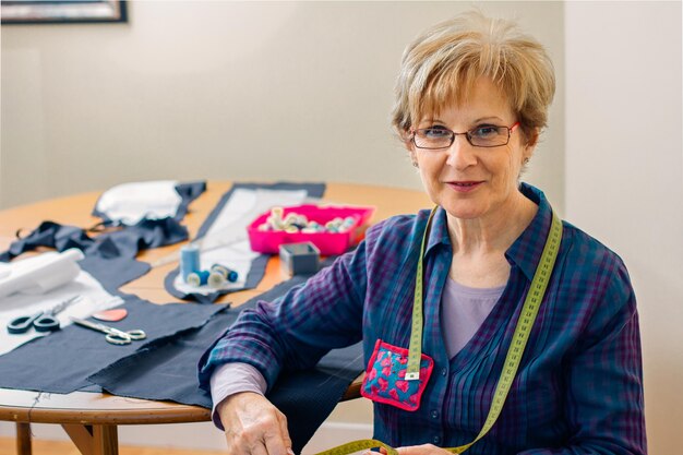 Costureira sênior posando olhando para a câmera com uma mesa com materiais de costura no fundo