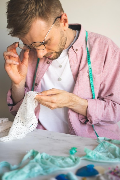 Un costurero autodidacta con una cinta métrica en el cuello Un diseñador masculino con gafas elige el tipo de encaje para la ropa interior Un sastre con una camisa rosa trabaja en casa hombre cosiendo lencería Estilo de vida