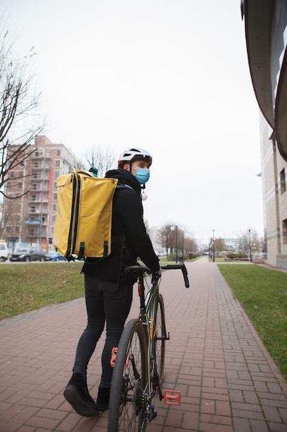 Courier com máscara médica, carregando mochila térmica, caminhando pela cidade de bicicleta