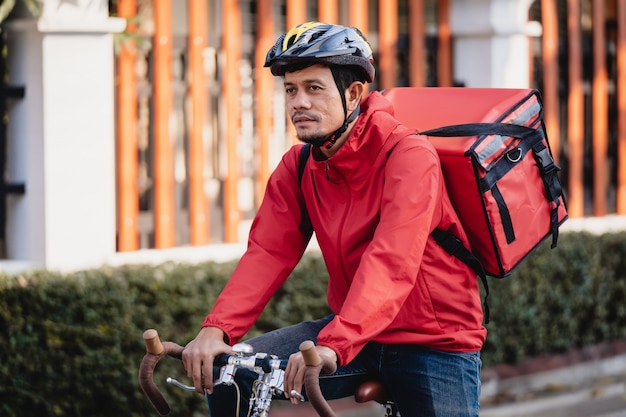 Courier de uniforme vermelho com caixa de entrega nas costas andando de bicicleta e olhando no celular para conferir o endereço para entregar comida ao cliente. Correio de bicicleta entregando comida na cidade.