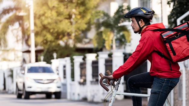 Courier de uniforme vermelho com caixa de entrega nas costas andando de bicicleta e olhando no celular para conferir o endereço para entregar comida ao cliente. correio de bicicleta entregando comida na cidade.