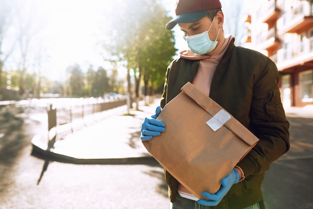 Courier em máscara protetora e luvas médicas em uma scooter entrega pacotes de papel ofício com alimentos. Serviço de entrega em quarentena, surto de doença, condições pandêmicas de coronavírus covid-19.