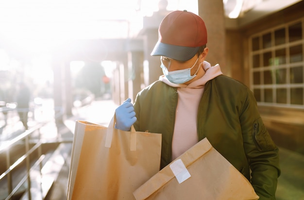 Courier em máscara protetora e luvas médicas em uma scooter entrega pacotes de papel ofício com alimentos. Serviço de entrega em quarentena, surto de doença, condições pandêmicas de coronavírus covid-19.