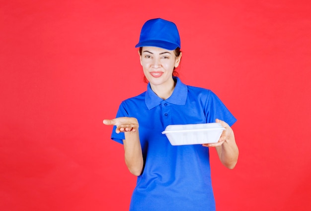 Courier mulher de uniforme azul segurando uma caixa para viagem e apontando para a pessoa à frente.