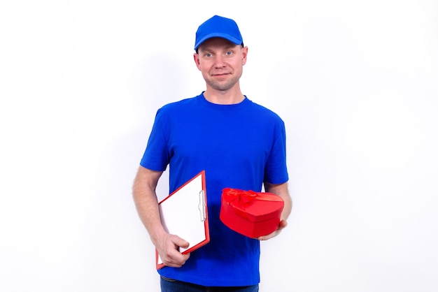 Courier en uniforme azul sostiene una caja de regalo roja en forma de corazón para el Día de San Valentín. Entrega a domicilio de