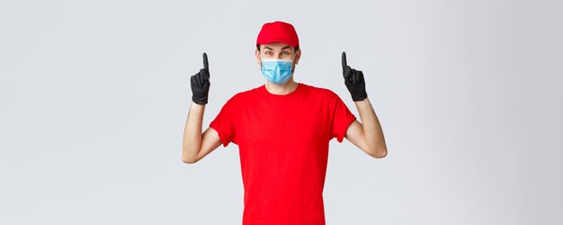 Covid-19, auto-quarentena, compras online e conceito de remessa. Entregador feliz divertido anunciar serviço de empresa de correio durante o bloqueio por coronavírus, apontando o dedo para cima, usar máscara médica.