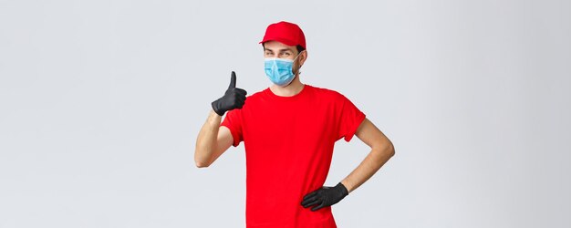 Covid-19, autocuarentena, concepto de compras y envío online. El amable y alegre repartidor con uniforme rojo, guantes y mascarilla garantiza la calidad del servicio de mensajería, muestra el pulgar hacia arriba en me gusta, aprobación.