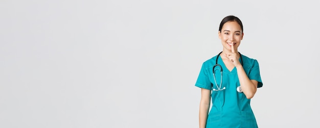 Covid-19, Beschäftigte im Gesundheitswesen, Pandemiekonzept. Fröhliche, lächelnde asiatische Krankenschwester in Scrubs, die ein Geheimnis haben, Überraschung machen, Psst-Geste zeigen, Pssst, ruhig bleiben, weißer Hintergrund stehen.
