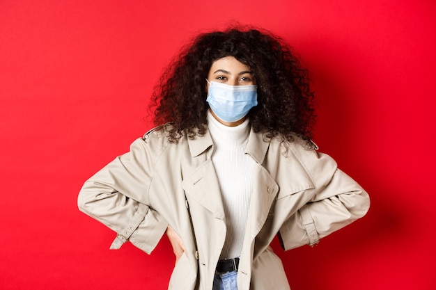 Covid-19, conceito de pandemia e quarentena. mulher moderna elegante com cabelo encaracolado, vestindo sobretudo e máscara médica, sorrindo para a câmera, fundo vermelho.